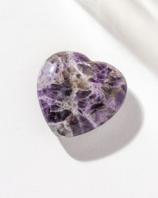 A heart-shaped Chevron Amethyst gemstone with a purple and white pattern, displayed on a light background.