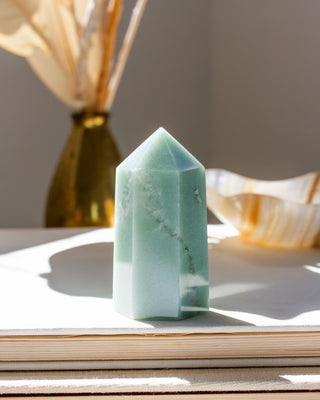 A green aventurine gemstone carved into a faceted tower shape, displayed on a flat stable with out of focus decorative items in the background.