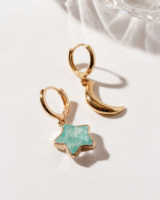 A pair of gold-colored huggie hoop earrings, one with a amazonite star-shaped gemstone and the other with a crescent moon shape, placed against a white surface.