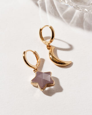 A pair of gold-colored huggie hoop earrings, one with a star-shaped coffee moonstone gemstone and the other with a gold crescent moon shape, placed against a reflective surface.