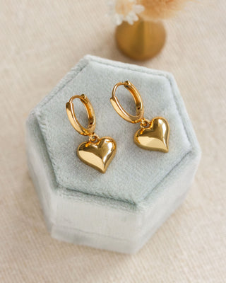 Gold heart-shaped earrings on a hexagonal green velvet stand.