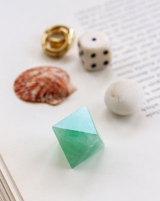 A carved green fluorite gemstone in the shape of an octahedron, displayed on a flat surface with a golden ring, a dice, and a seashell in the background.