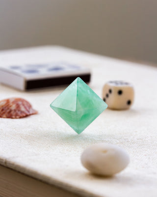 A carved green fluorite gemstone in the shape of an octahedron, displayed on a flat surface with a dice, and a seashell in the background.