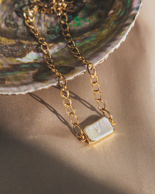 A minimalist collar chain necklace with a rectangular pearl centerpiece, dangling from a shell on a beige fabric surface.