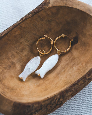 Gold hoop earrings with fish-shaped moonstone charms on a wooden surface.