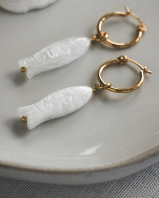 Gold hoop earrings with white moonstone fish-shaped pendants on a light gray plate.