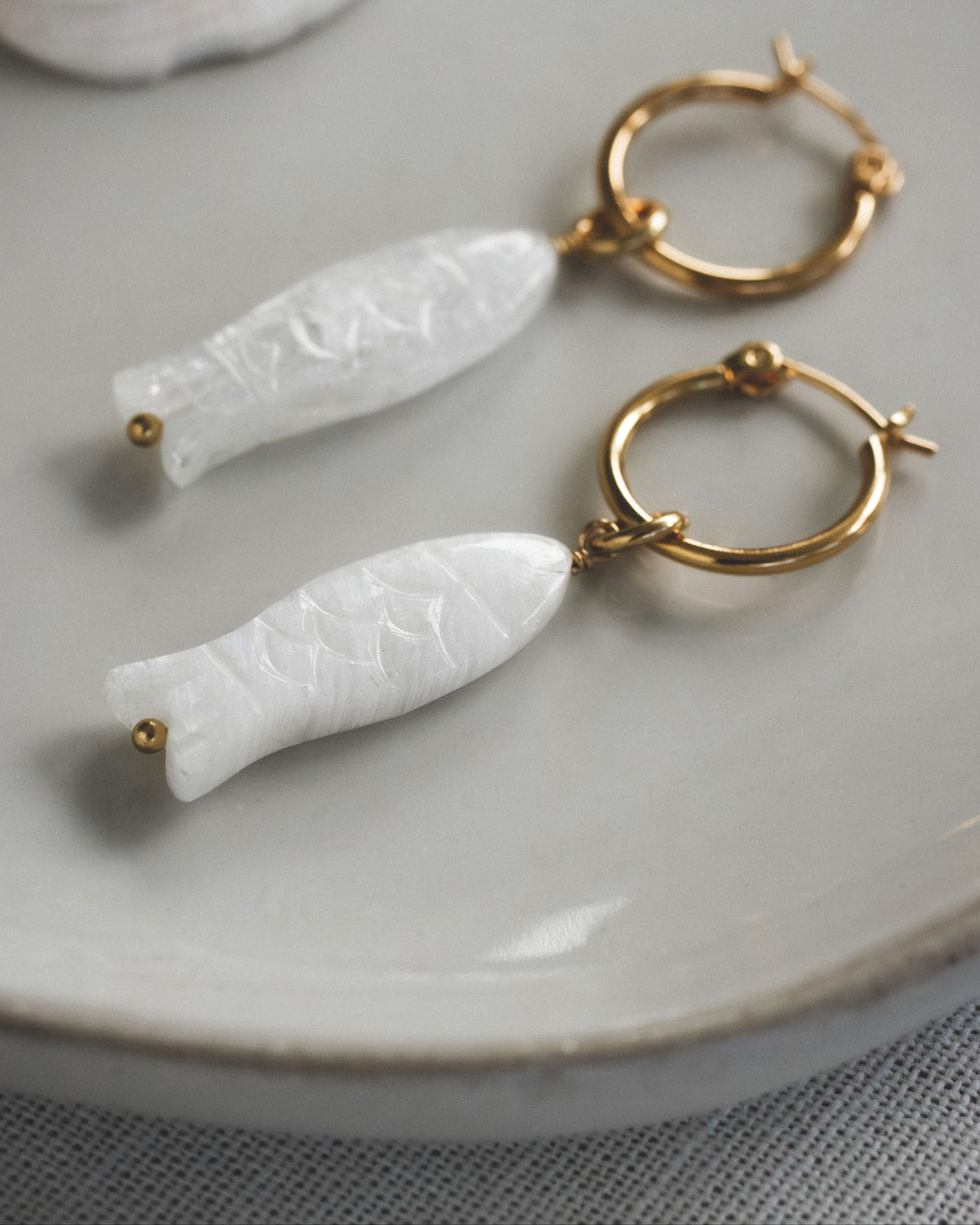 Gold hoop earrings with white moonstone fish-shaped pendants on a light gray plate.