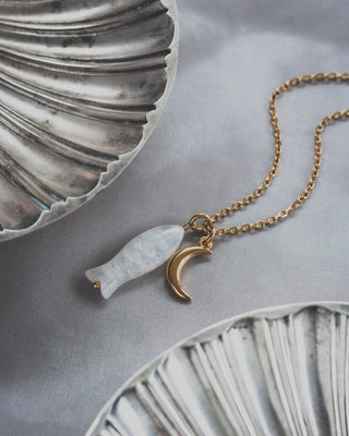 Necklace with moonstone fish and gold moon pendant near decorative shell plates on a gray background.