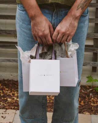 A person wearing a green sweater and light-wash denim jeans holds two white Luna Norte shopping bags with purple ribbon handles. The bags are filled with white and black polka-dotted tissue paper, suggesting a gift.