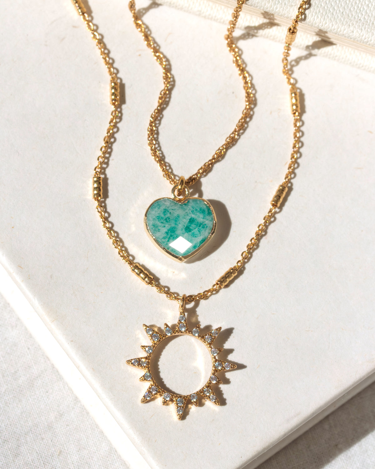 Two gold necklaces with amazonite heart and sun pendants on a white surface