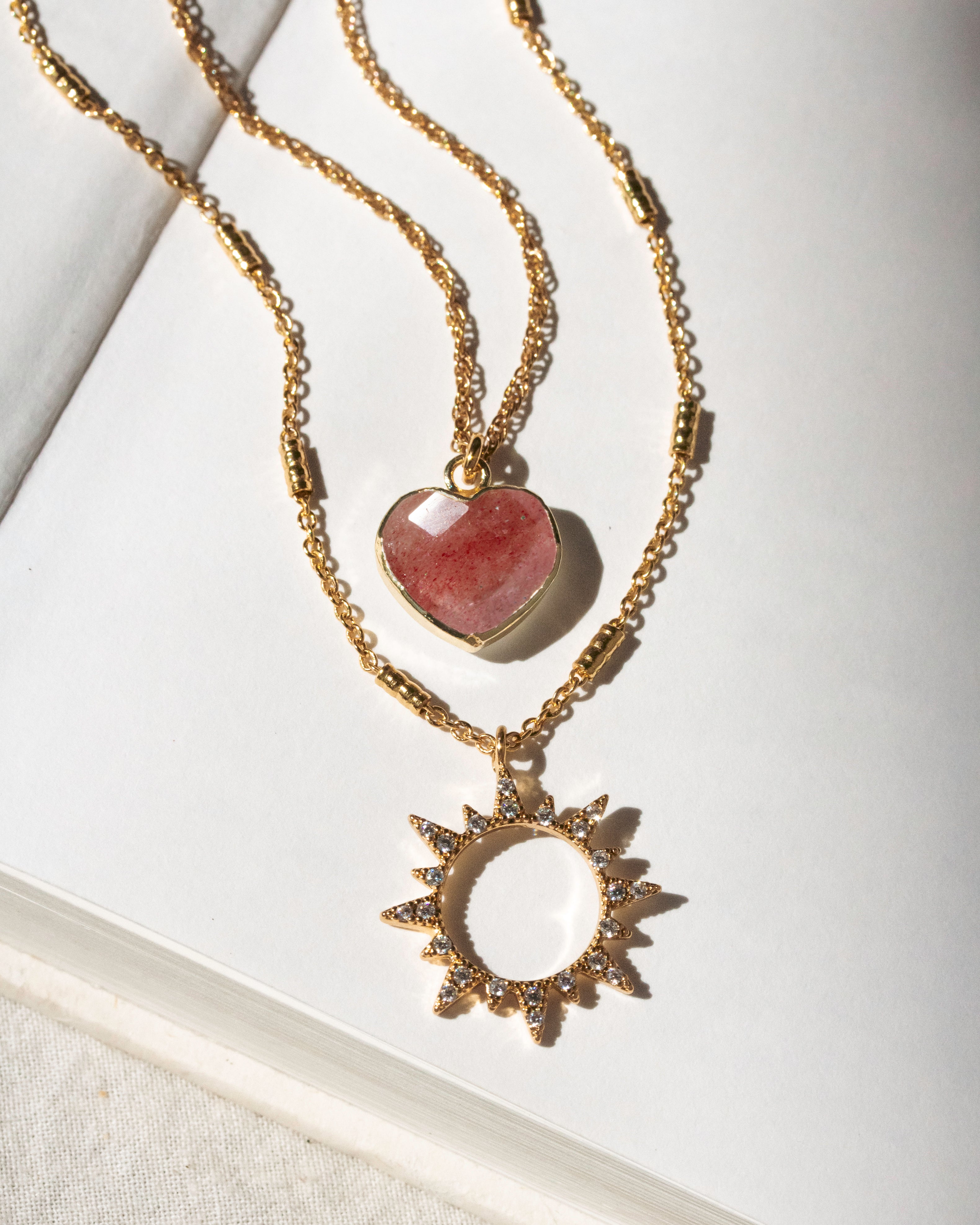 Gold necklaces with strawberry quartz heart and sun-shaped pendants on a white background