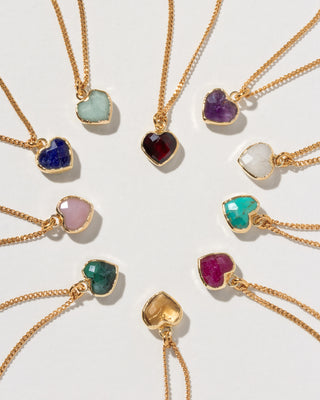 Nine gold heart-shaped pendant necklaces with different colored gemstones are arranged in a heart on a light gray background.