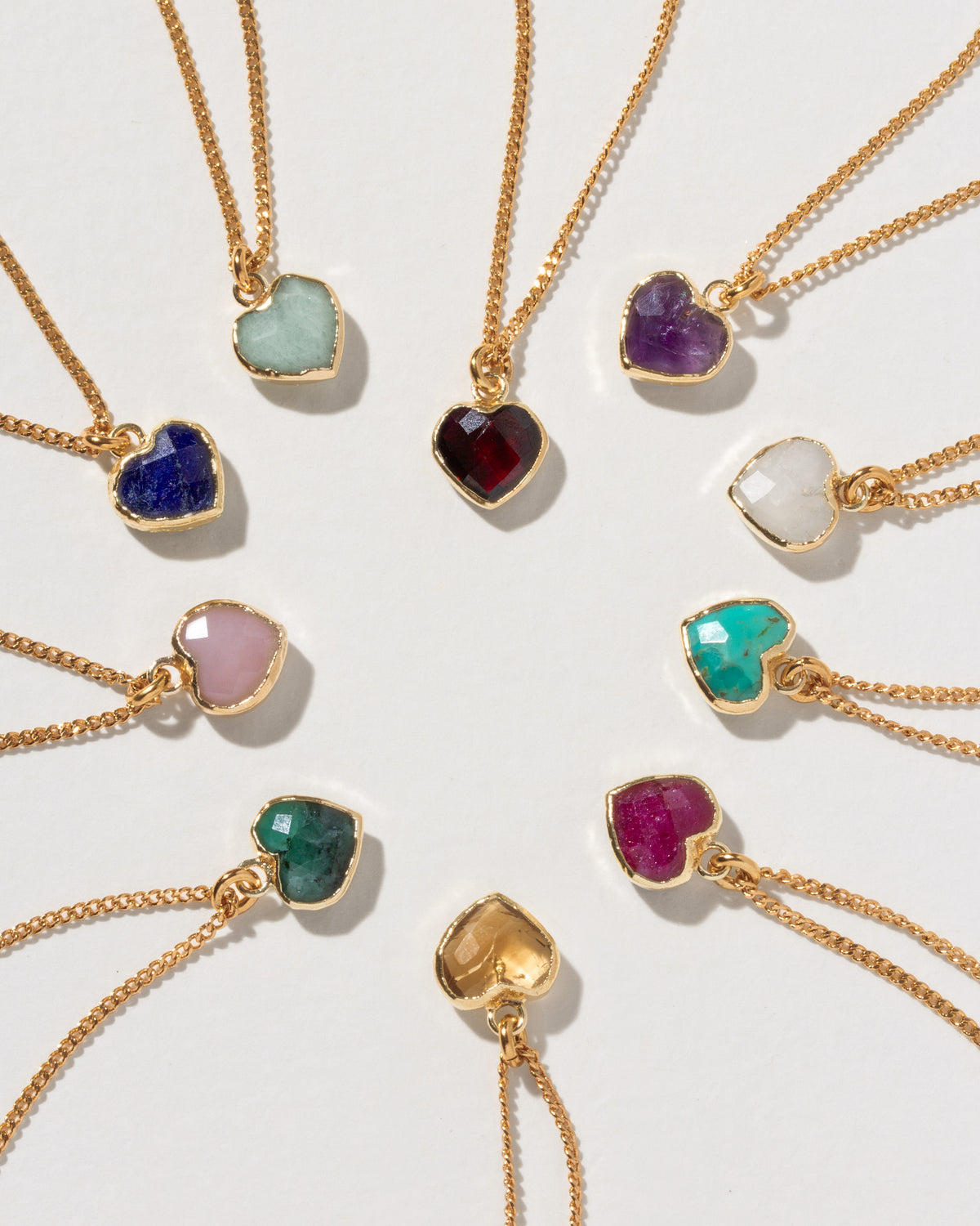 Nine gold heart-shaped pendant necklaces with different colored gemstones are arranged in a heart on a light gray background.