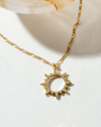 Gold "Caffeine" necklace with a sunburst pendant on a white surface.