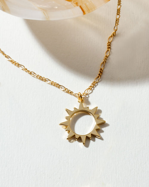 Gold "Caffeine" necklace with a sunburst pendant on a white surface.