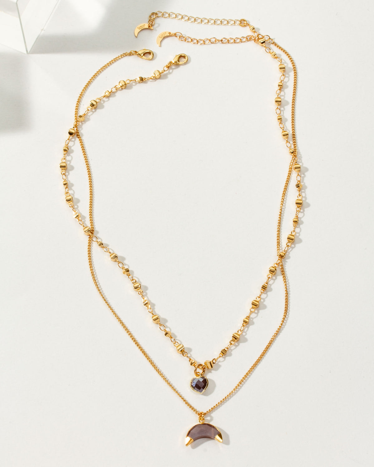 A layered, golden necklace with a heart charm and a coffee moonstone crescent moon charm.