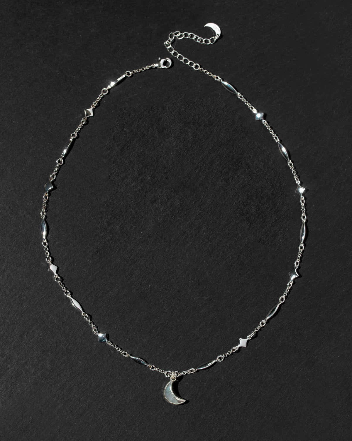 A silver-toned chain necklace featuring small geometric star and diamond-shaped stations, a matching crescent moon pendant, and an adjustable extender chain.