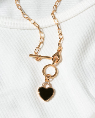 A  gold chain necklace with a toggle clasp and a heart-shaped pendant featuring a glossy black onyx center, displayed on a white ribbed knit top.