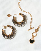 A gold-toned jewelry set on a white textured background, featuring a pair of textured hoop earrings, a matching chain necklace with a heart charm.