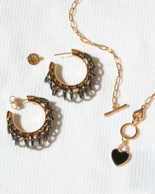 A gold-toned jewelry set on a white textured background, featuring a pair of textured hoop earrings, a matching chain necklace with a heart charm.