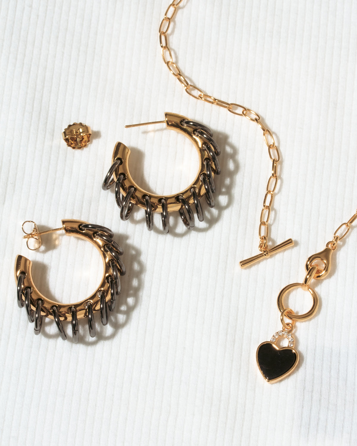 A gold-toned jewelry set on a white textured background, featuring a pair of textured hoop earrings, a matching chain necklace with a heart charm.