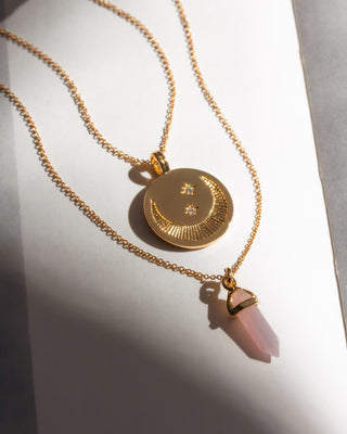 Gold necklace with a circular pendant and pink crystal charm on a white background