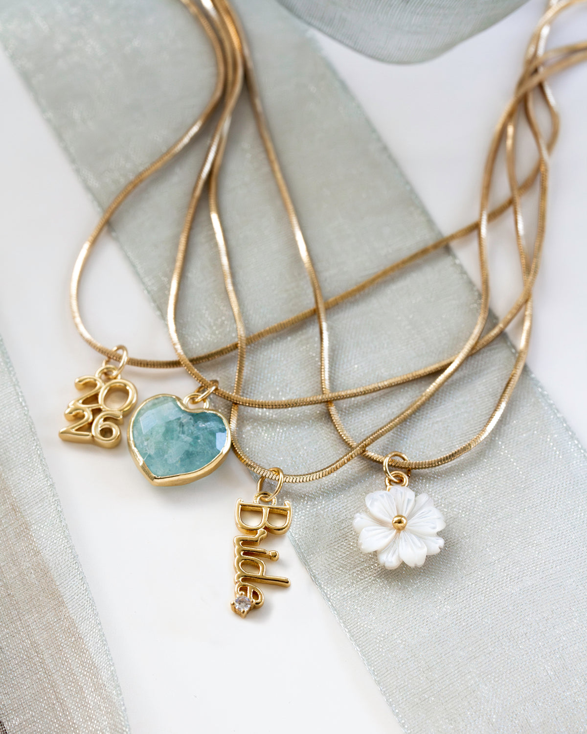 The necklaces include charms: a gold 2026 script pendant, a light blue heart-shaped stone pendant, a white flower charm, and a gold Bride lettering charm.