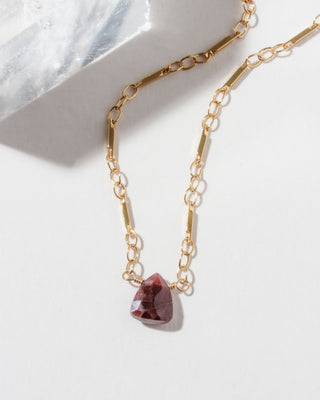 Closeup of gold necklace with a triangular red garnet gemstone pendant on a white background