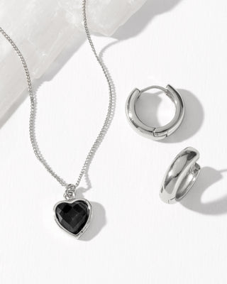 Silver jewelry set featuring a heart-shaped pendant necklace with a black onyx heart inlay, and matching silver huggie hoop earring.