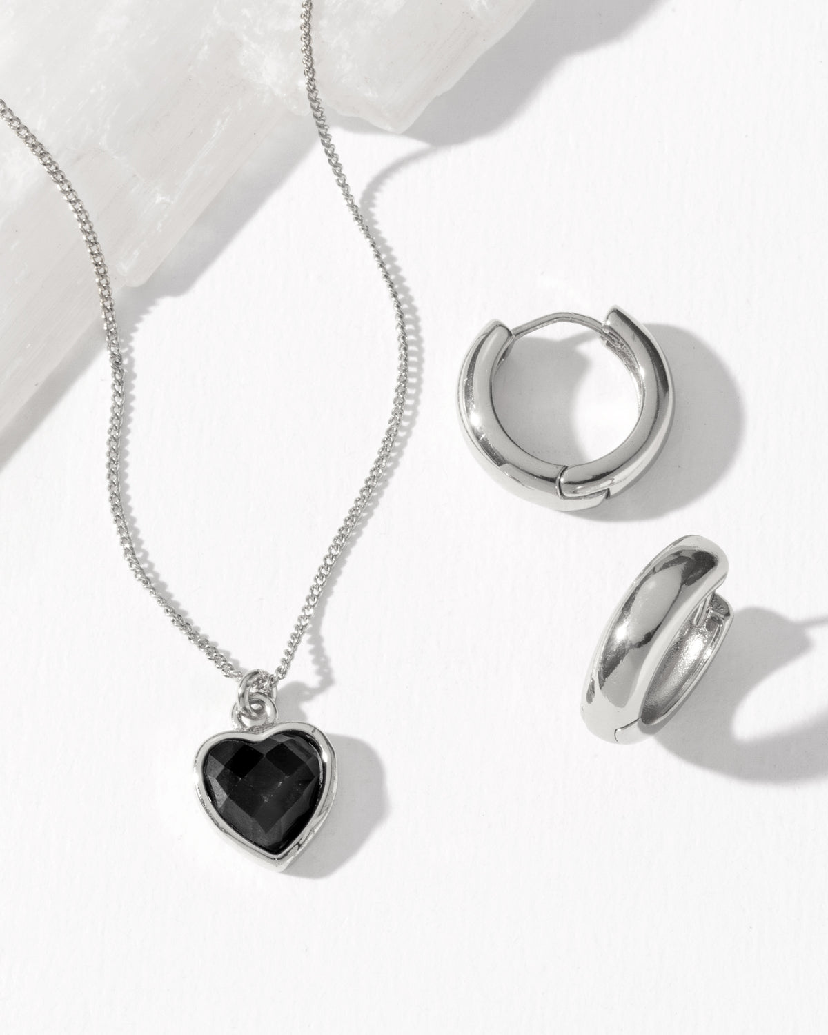 Silver jewelry set featuring a heart-shaped pendant necklace with a black onyx heart inlay, and matching silver huggie hoop earring.