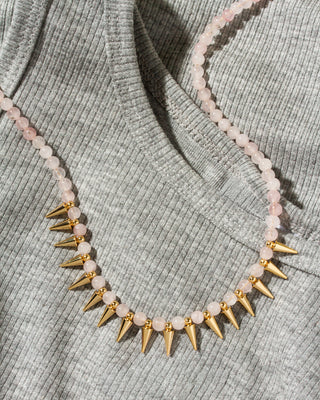 A close-up of a necklace draped over a ribbed gray knit top. The necklace features pale pink round beads with alternating gold spikes.