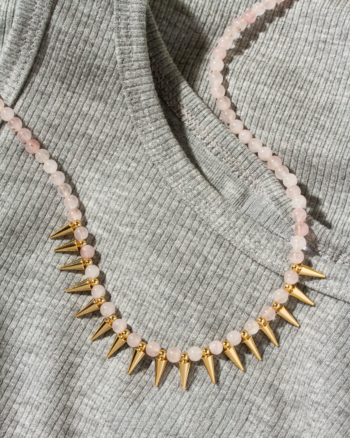 A close-up of a necklace draped over a ribbed gray knit top. The necklace features pale pink round beads with alternating gold spikes.