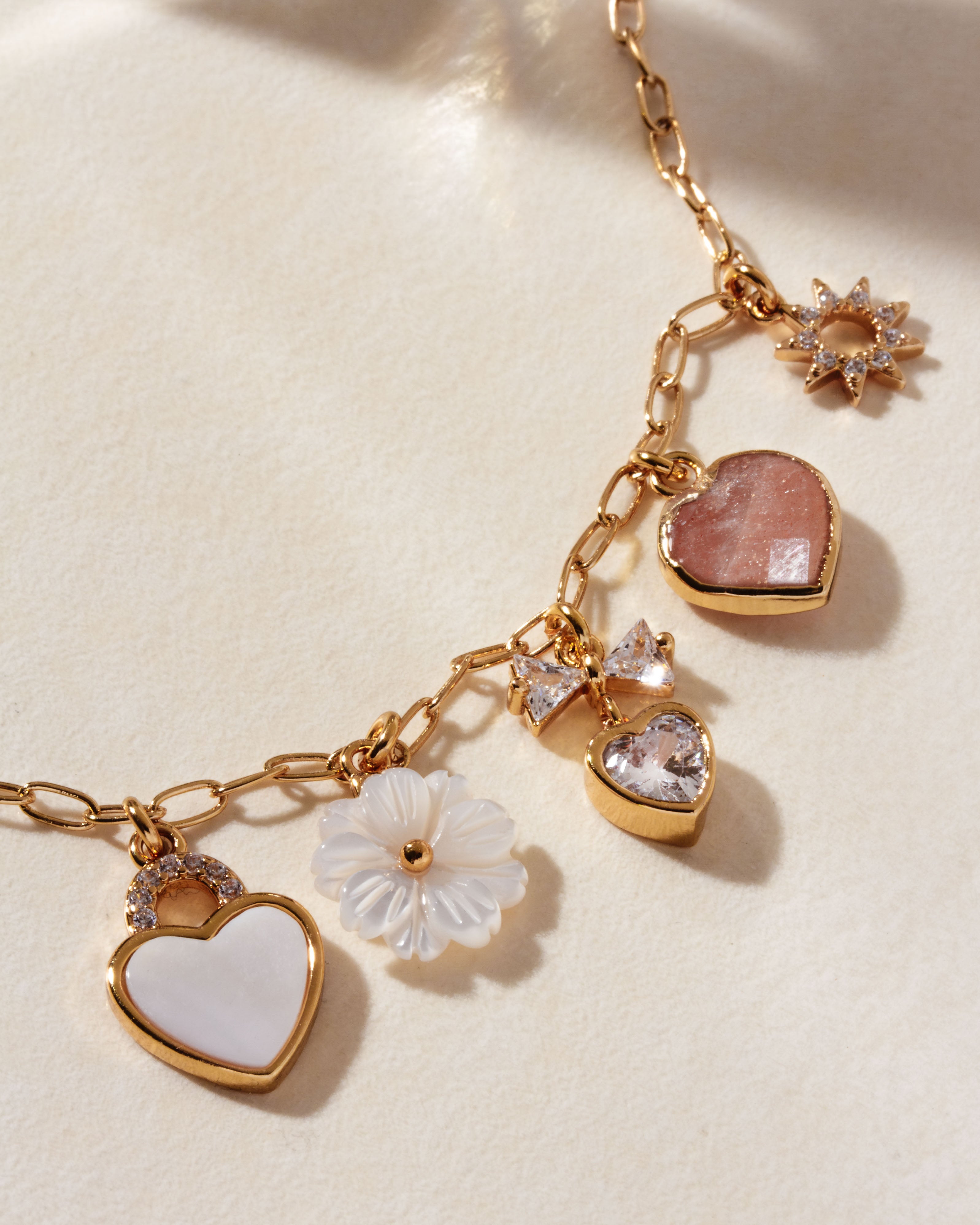 Gold charm necklace with mother-of-pearl and rose quartz hearts, and other floral and star charms.