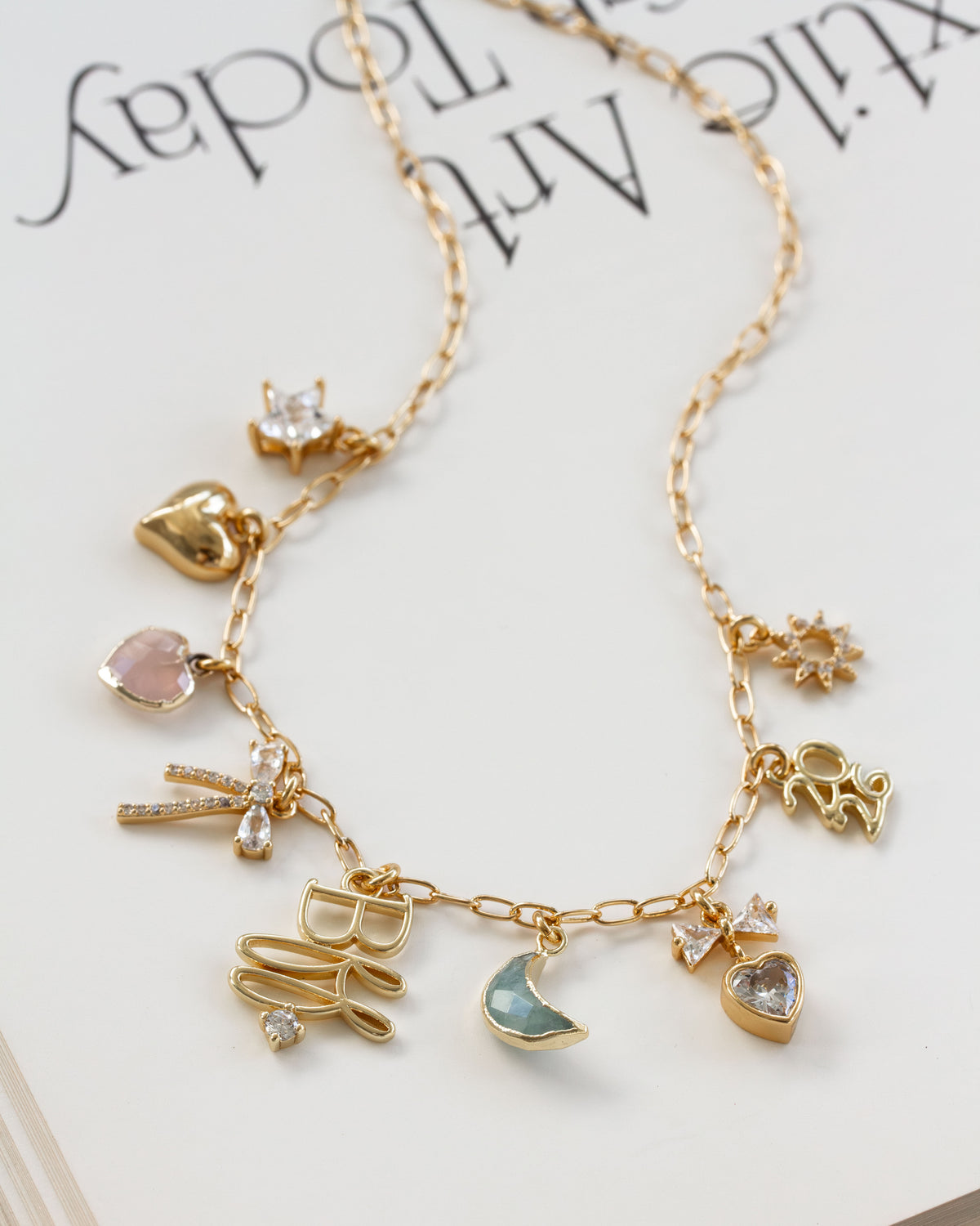 A gold-tone charm necklace  features multiple delicate charms, including a bow, moon, star, and hearts, some embellished with clear rhinestones and pastel-colored stones.