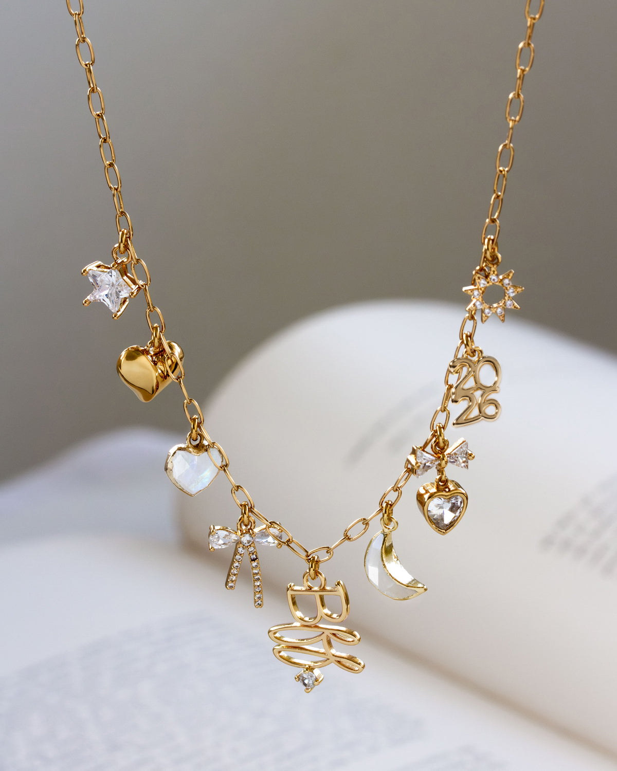 A gold-tone charm necklace with multiple delicate charms including hearts, a crescent moon, stars, and a bow, displayed on an open book against a soft gray background.