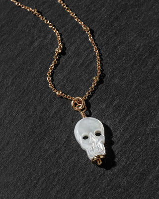 A gold chain necklace featuring a carved white skull pendant, displayed on a dark textured slate background.