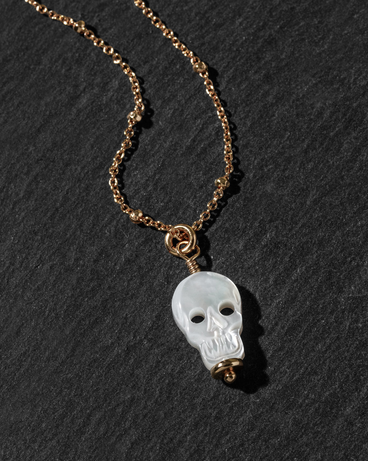 A gold chain necklace featuring a carved white skull pendant, displayed on a dark textured slate background.