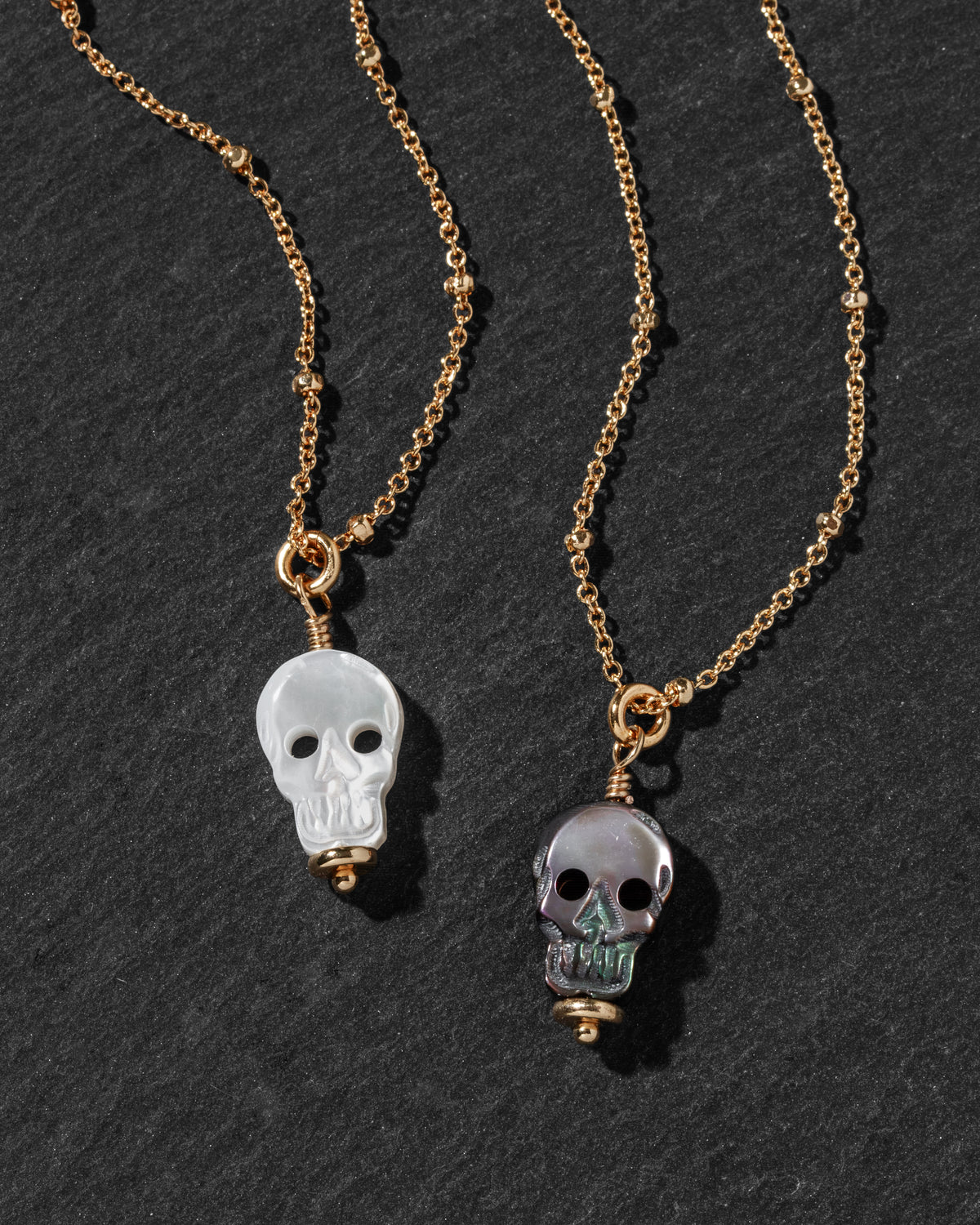 Two gold chain necklaces with skull pendants, one featuring a translucent white skull and the other a translucent gray skull.
