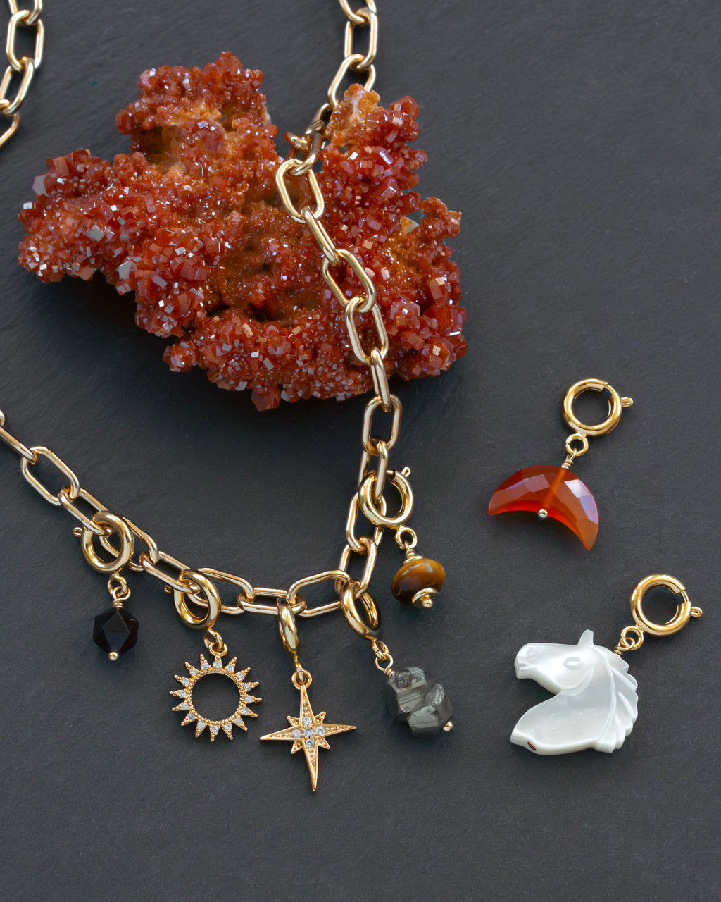 Gold charm necklace with various charms on a dark surface with a red crystal rock.
