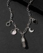A silver-toned chain necklace with multiple charms, including a sun, moon, heart, and a faceted crystal pendant, displayed on a dark textured surface.