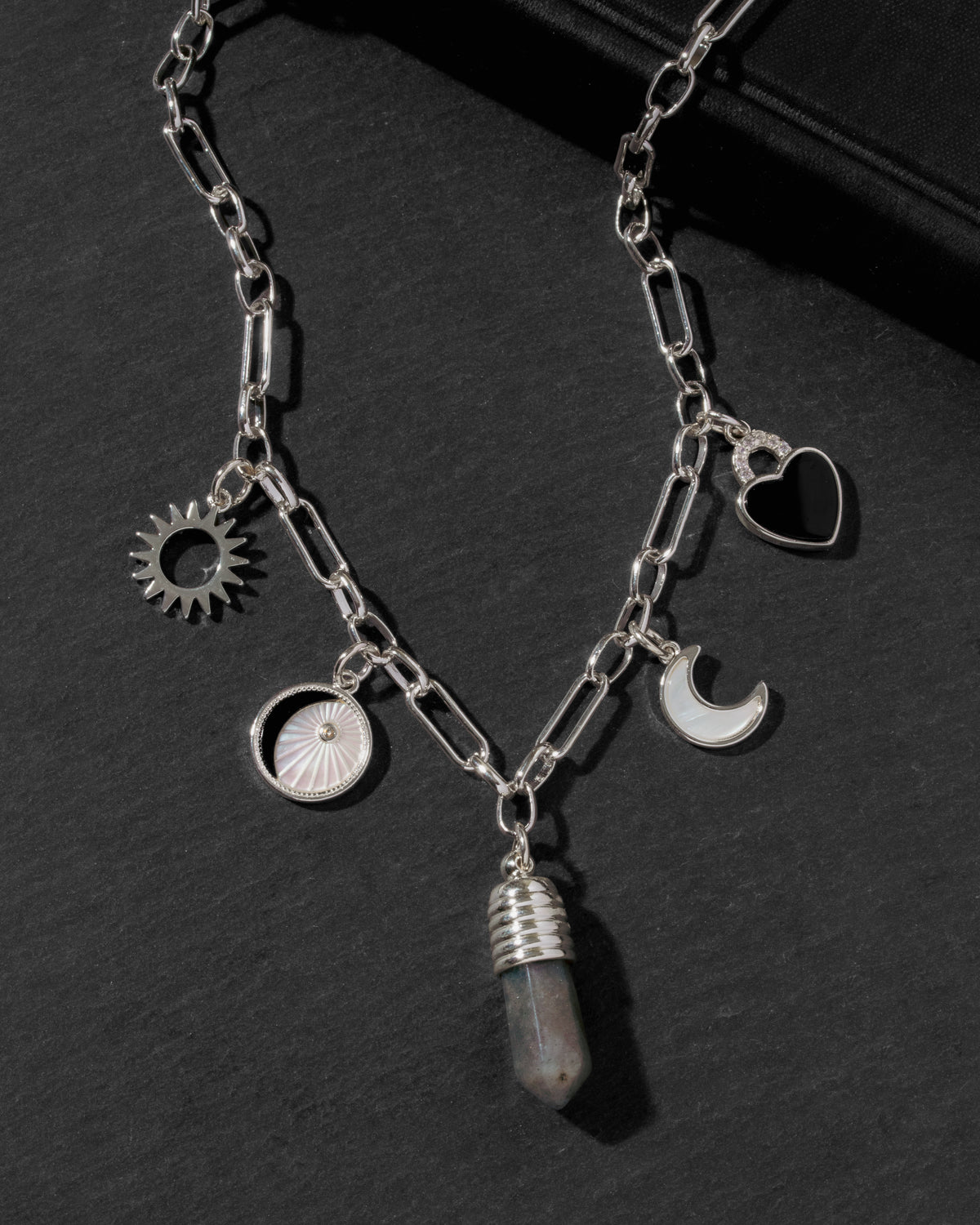 A silver-toned chain necklace with multiple charms, including a sun, moon, heart, and a faceted crystal pendant, displayed on a dark textured surface.