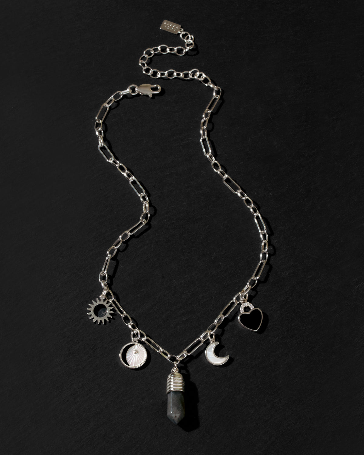 A silver-toned chain necklace with multiple charms, including a sun, a heart, a crescent moon, a moon phase, and a labradorite crystal pendant, displayed on a dark textured background.