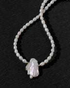 A beaded necklace featuring irregular white rice pearls and a large, organic-shaped white pearl pendant, accented with small gold spacers, displayed on a dark textured background.