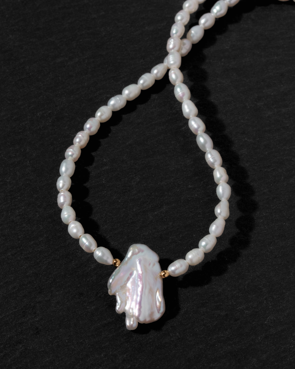 A beaded necklace featuring irregular white rice pearls and a large, organic-shaped white pearl pendant, accented with small gold spacers, displayed on a dark textured background.