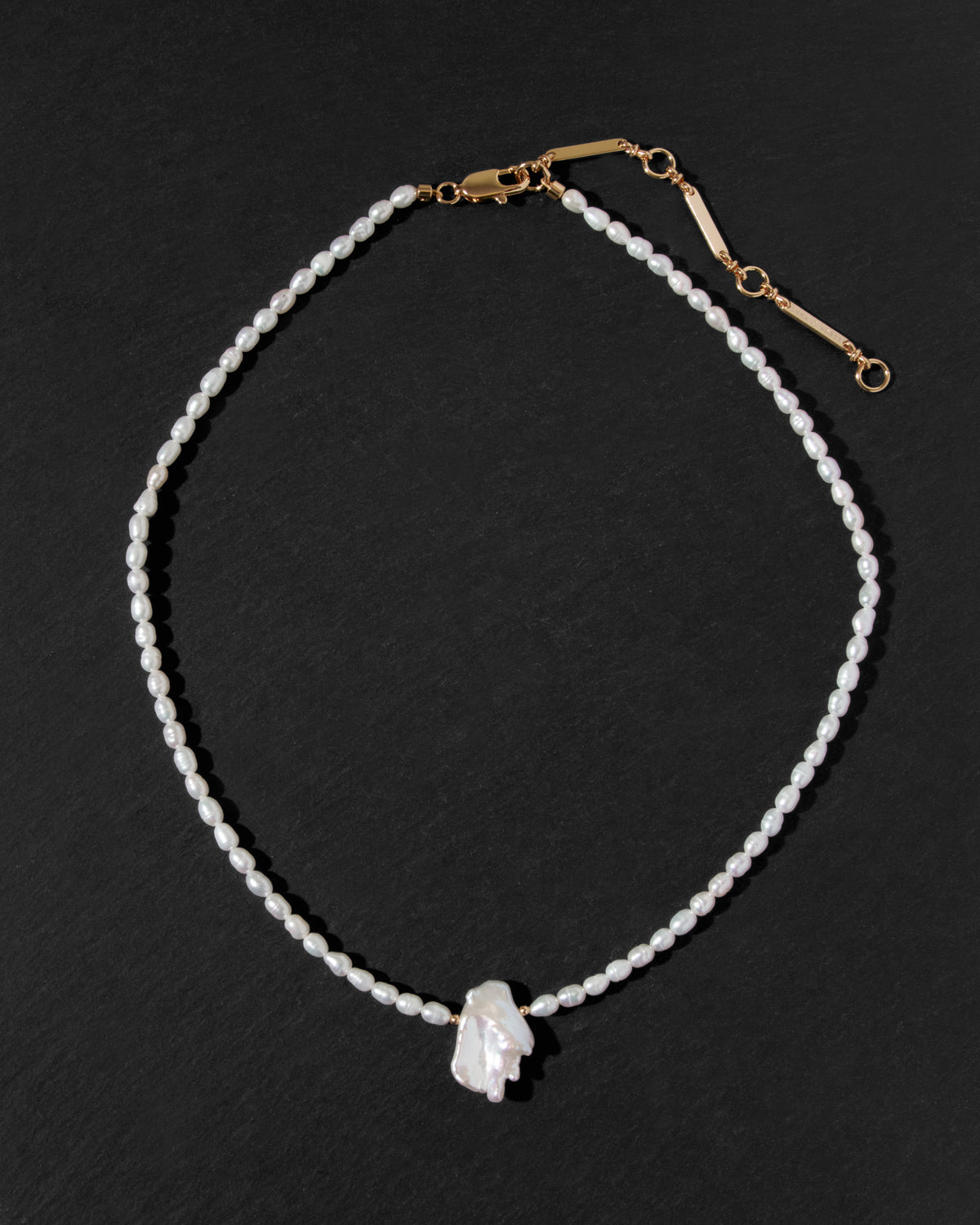A beaded classic necklace featuring a single strand of small white rice pearls with a central white keshi pearl pendant and gold-tone hardware, displayed on a dark textured background.