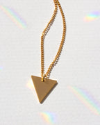 Gold triangle pendant necklace on a light gray background with rainbow shadows