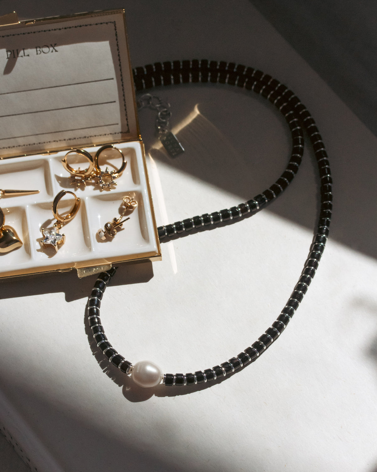 Black beaded necklace with a pearl pendant next to an open jewelry box on a reflective surface.