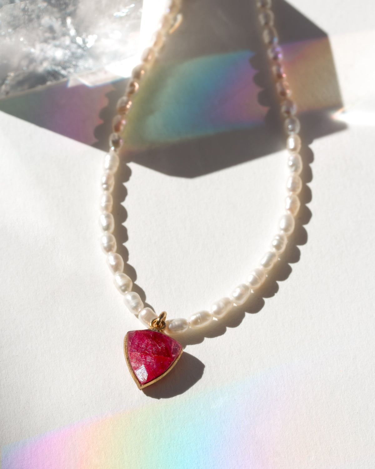 Necklace with a inverted triangle-shaped pink pendant on a white surface with rainbow reflection