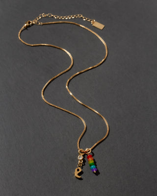 Gold necklace with rainbow charm and letter 'e' initial charm on a dark gray background