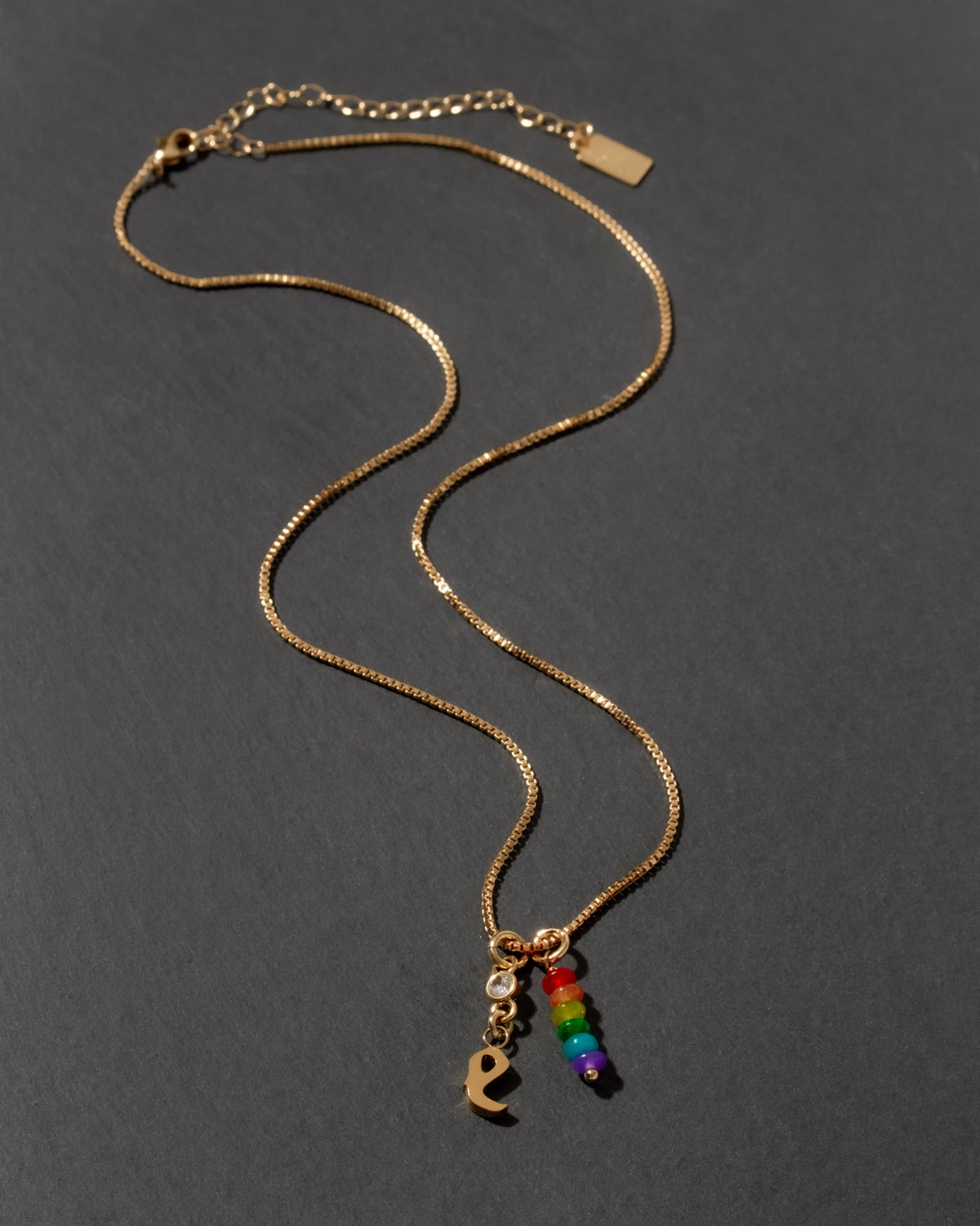 Gold necklace with rainbow charm and letter 'e' initial charm on a dark gray background